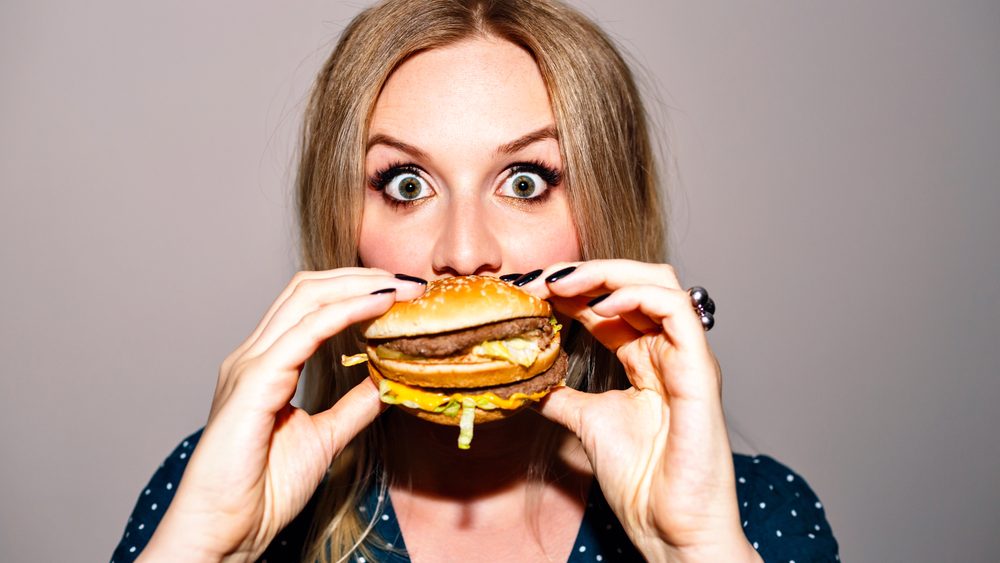 Close up funny crazy image of blonde pretty woman enjoying her big tasty burger, fast junky unhealthy food, image with flash. Big surprising eyes, holding burger near face.
