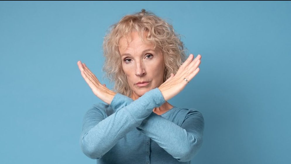Serious mature woman showing stop gesture with crossed hands. Sve the social distance. Studio shot on blue wall.