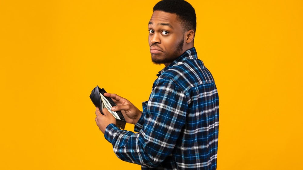 Low Salary. Discontented Black Man Holding Wallet With Cash Money Looking At Camera Standing On Yellow Studio Background.