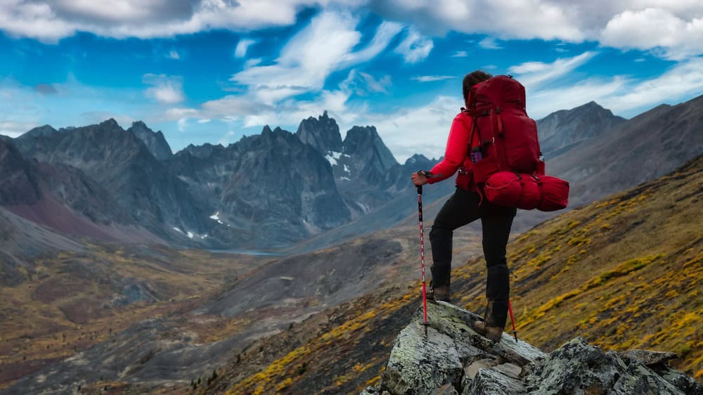 Woman Standing on Rocks looking at Scenic Mountain Peaks and Valley during Fall in Canadian Nature. Colorful Cloudy Sky Artistic Render. Landscape in Tombstone Territorial Park, Yukon, Canada.