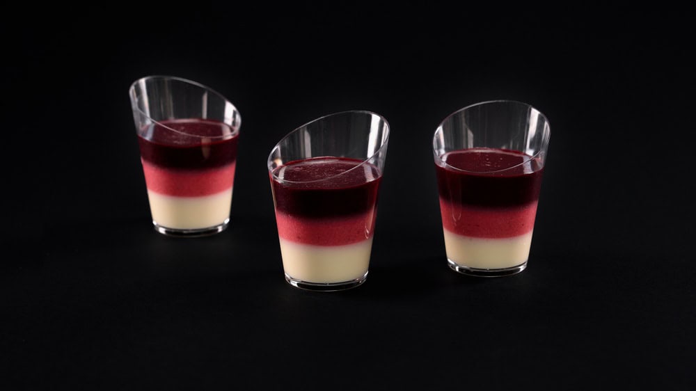 Jelly sweet dessert in three glasses, white milky, red and pink layers isolated on black background. Front closeup view of fresh served delicious mousse. Concept of sweet food.