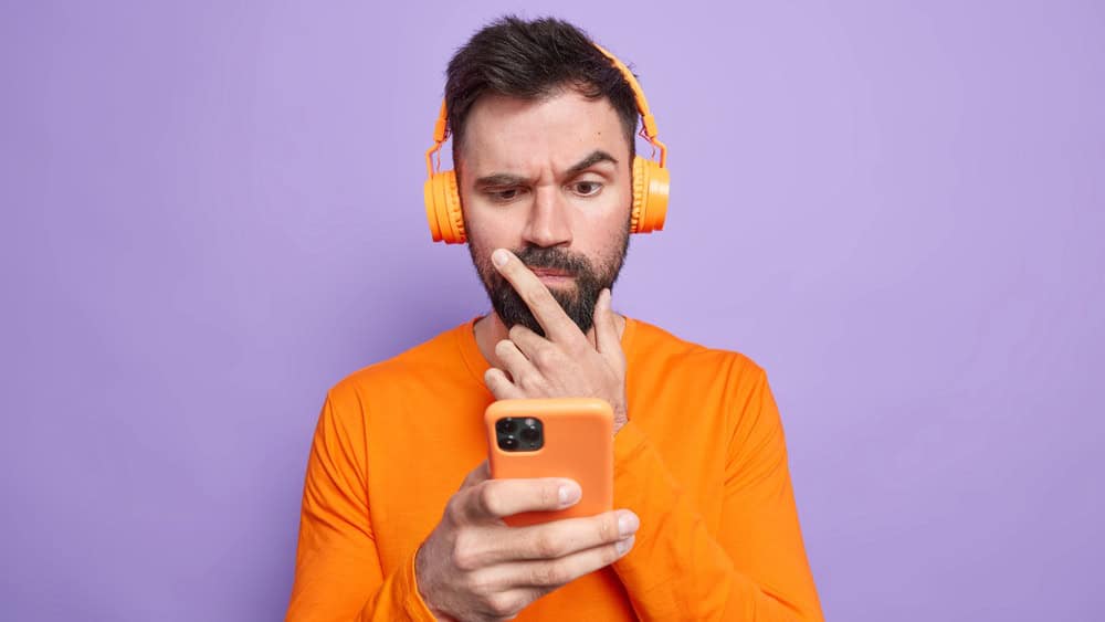 Puzzled displeased man looks attentively at smartphone display uses mobile phone for checking news online wears headphones on ears poses alone against purple background. Lifestyle technology