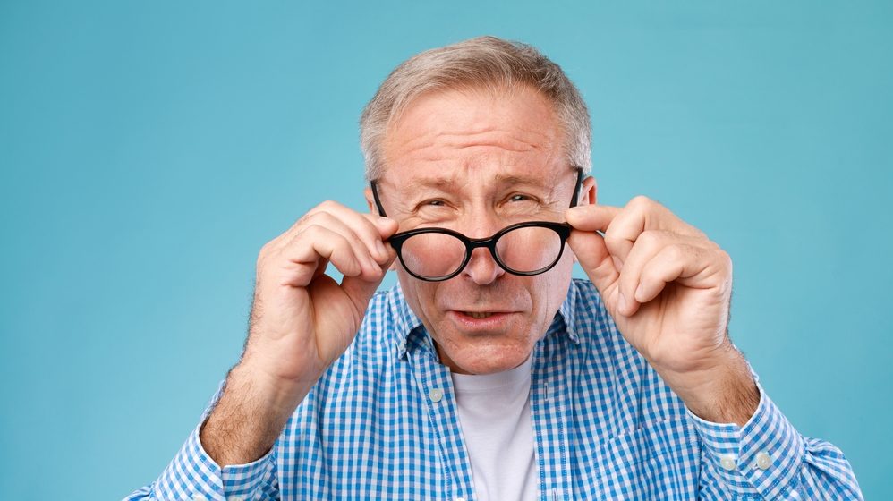 Poor Eyesight. Senior Man Can't See, Squinting Eyes Wearing Glasses Having Problems With Vision, Looking At Camera At Blue Studio. Ophtalmic Issue, Bad Sight In Older Age, Macular Degeneration Concept