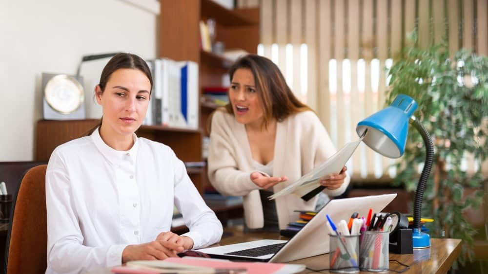 Angry business woman criticizing sad frustrated female employee sitting at office desk at work