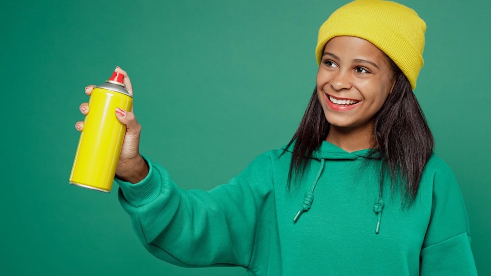 Little kid teen girl of African American ethnicity 13-14 years old wear casual hoody hat hold aerosol spray paint in bottle for graffiti look aside isolated on plain green background Childhood concept