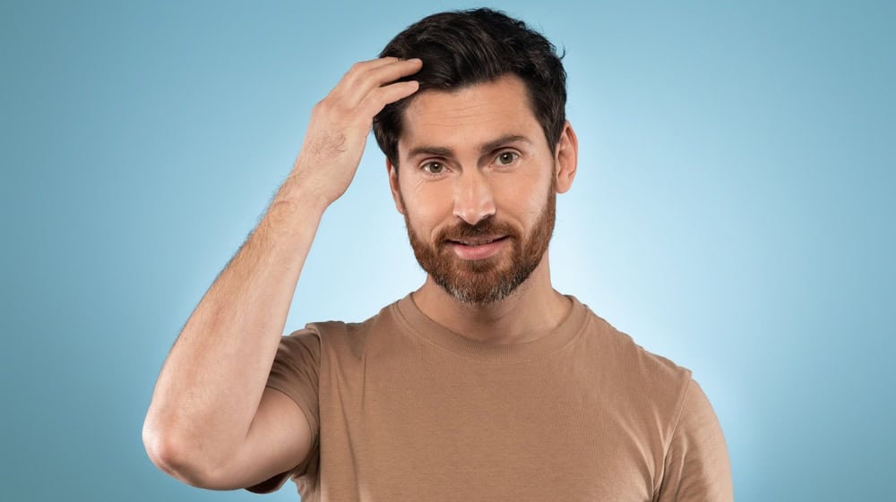 Handsome bearded middle aged man in t-shirt touching his hair while posing on blue studio background and smiling at camera, closeup portrait, copy space