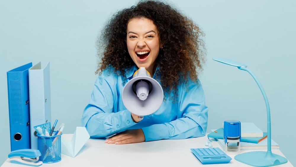Young excited employee business woman wears casual shirt sit work at white office desk scream in megaphone announces discounts sale Hurry up isolated on plain pastel blue background studio portrait