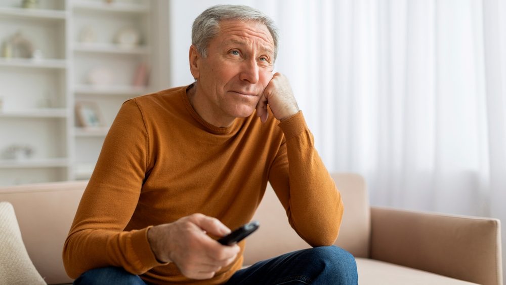 Unhappy senior man with remote control in his hand sitting on couch at home, feeling bored, looking for something nice interesting to watch on TV at weekend, copy space