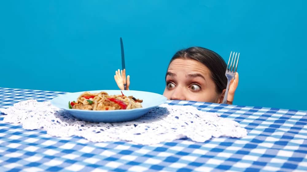 Young woman peeking over table, gazing at plate of noodles with toy hand holding knife and fork against blue background. Concept of food pop art photography, creativity, quirky style