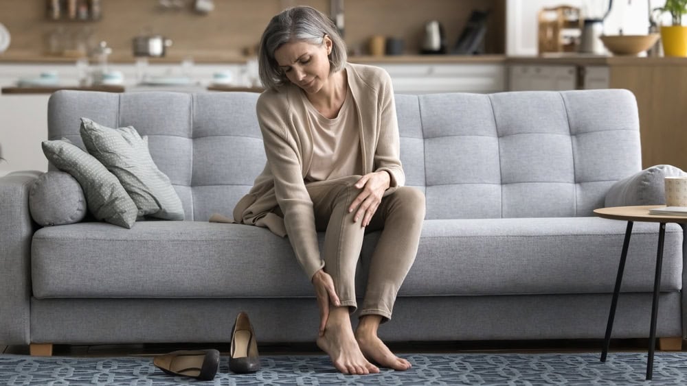 Female came home sit on sofa took off shoes, touch ankle doing self-massage of tired feet, suffers from varicose, painful feelings discomfort after long workday on heels, need rest, orthopedic insoles