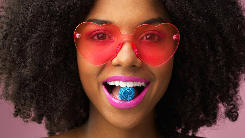 Portrait, candy and woman in studio with retro fashion, heart glasses or afro on pink background. Quirky, makeup and female person with bright lips, smile or fun eyewear for cosmetics or accessories