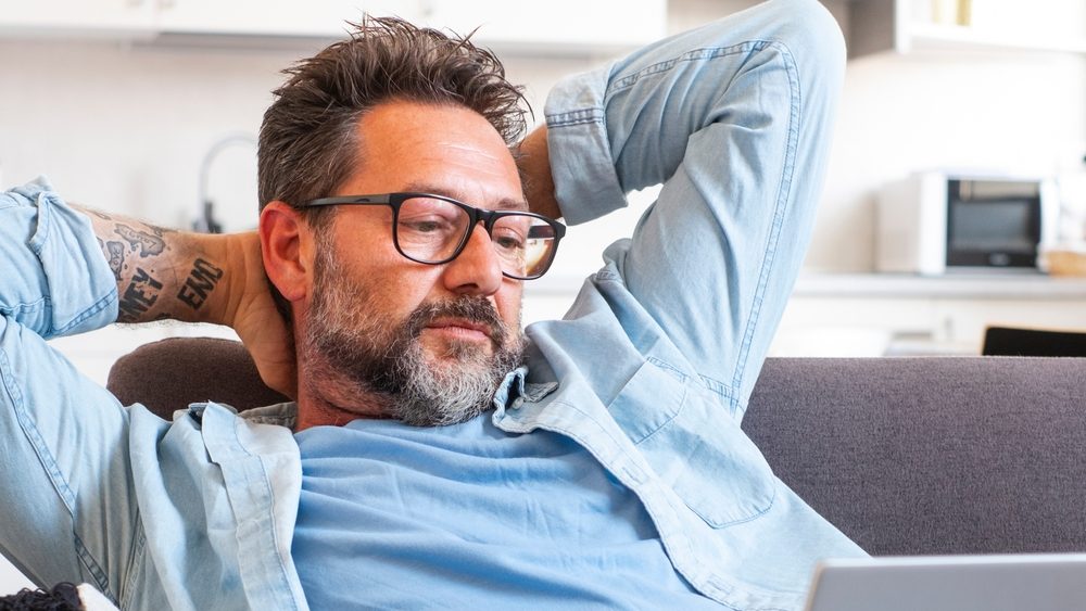 One adult man outstretching arms looking an open laptop sitting comfy on the couch at home. End work or browsing internet lazy afternoon concep tlifestyle people. Modern worker indoor apartment.