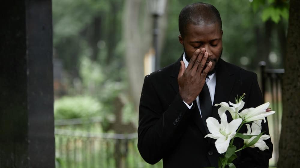 Medium shot of African American man in funeral black suit grieving at graveyard wiping tears and holding white lilies on sorrowful rainy day, copy space