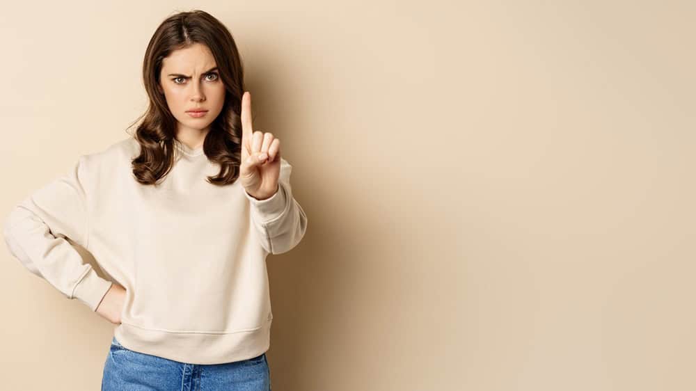 Serious woman show one finger, stop disapproval sign, forbid smth, say no, prohibit, standing over beige background.