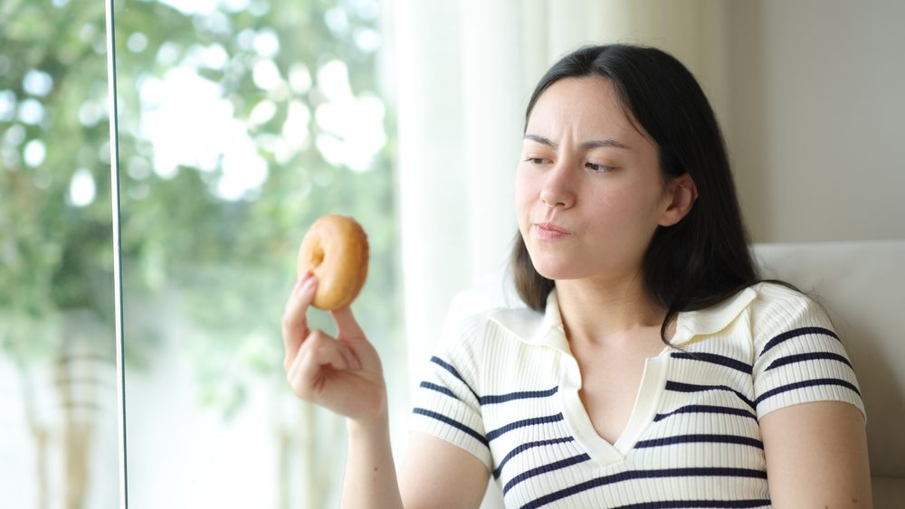 Doubtful asian woman looking at doughnut thinking if she should eat at home