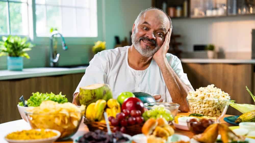 Senior Indian man sitting with table full of snacks, Asian foodie old man expressing happiness, joyful elderly person reacting to delicious food spread at modern home kitchen with excitement