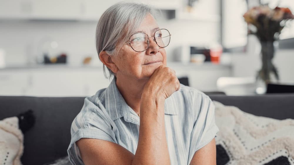 Sad elderly woman sits alone on a sofa at home, looking out the window with a worried expression. She reflects on challenges and feels stressed, illustrating loneliness and concern.