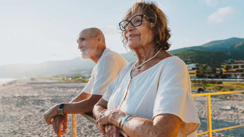 Elder man and woman standing together on a bright yellow lifeguard tower, looking out at the sunny beach and enjoying their active retirement lifestyle