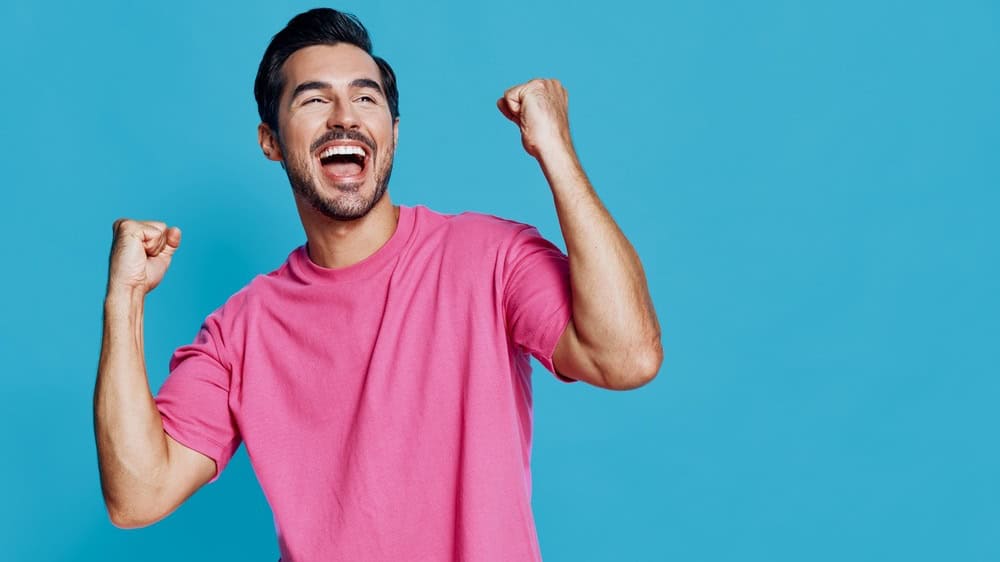 Full body joyful cheerful young man wearing pink t-shirt celebrating success with raised hands against a bright blue background. Positive emotions concept.