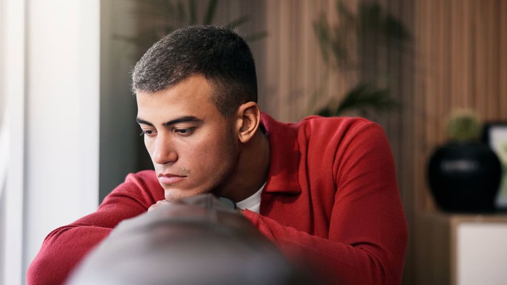 Sad, loneliness and man by window in home with ideas, mental health or anxiety on weekend break. Disappointed, upset and male person with depression, reflection or thinking in living room at house.