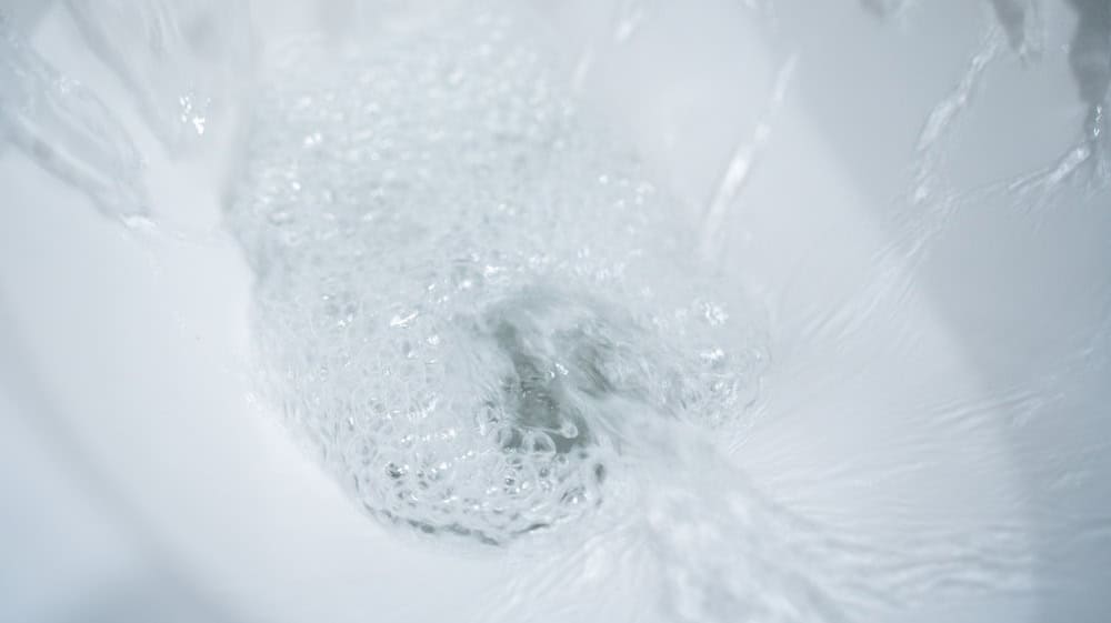 Water swirling in a white ceramic toilet bowl during a flush, foamy bubbles and motion blur forming a vortex around the drain, clean bathroom close up with bright highlights and minimal white interior