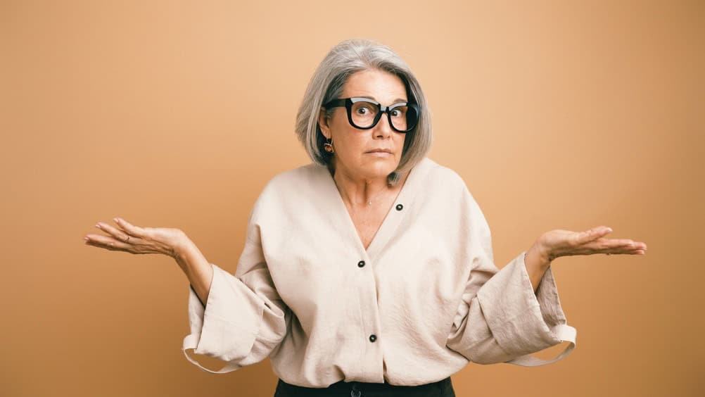 Senior woman shrugging, having a confused and uncertain expression, standing against an orange background