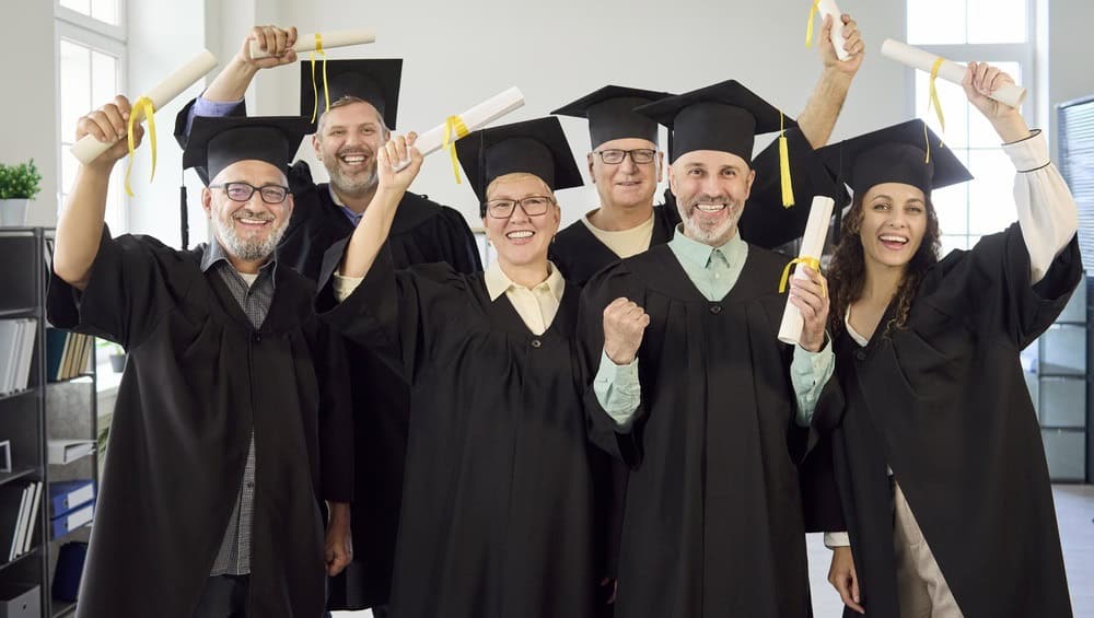 Adult graduates group celebrate graduation success with diplomas. Mature students in caps and gowns, smiling indoors, holding rolled certificates in a room. Simple clear concept of success.