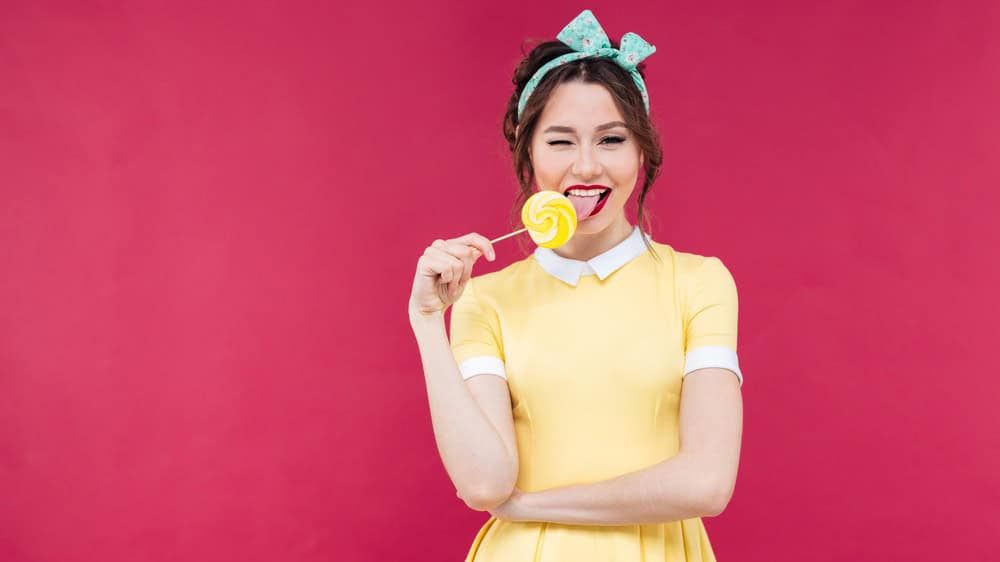 Happy charming pinup girl eating sweet lollipop and winking over pink background