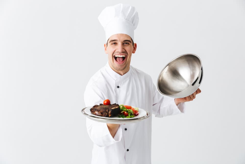 Excited man chef cook wearing uniform opening cloche cover isolated over white background, showing meat dish
