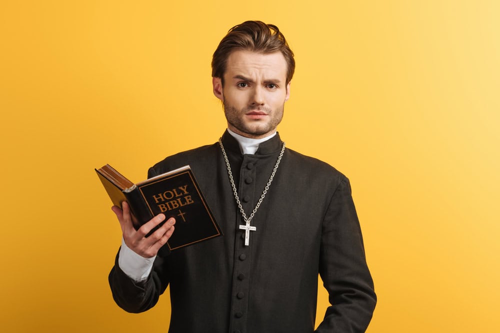 Surprised Catholic priest holding a Bible, looking at camera, isolated on a yellow background