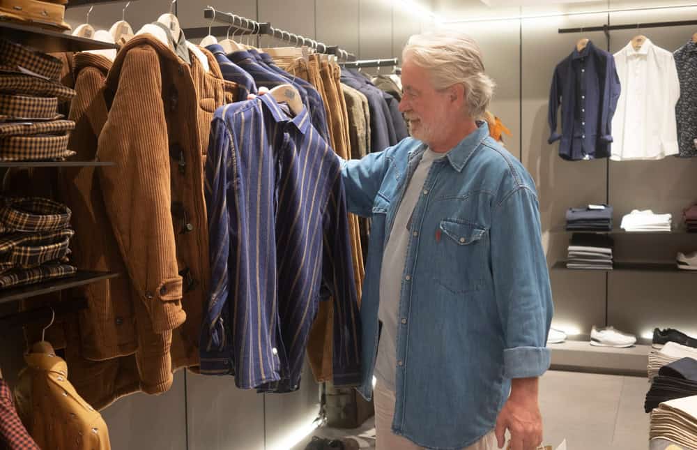One attractive senior man with a beard and white hair looks at the objects displayed in the store. His attention is turned to a striped shirt