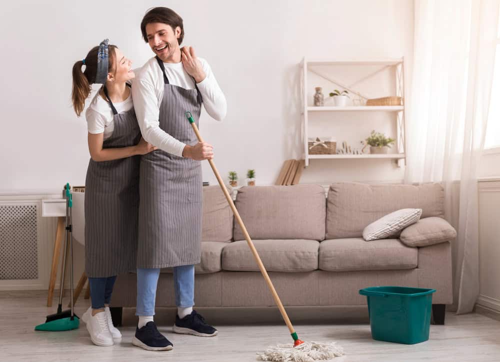Joyful young couple doing spring-cleaning together, mopping floor and hugging while tidying their apartment