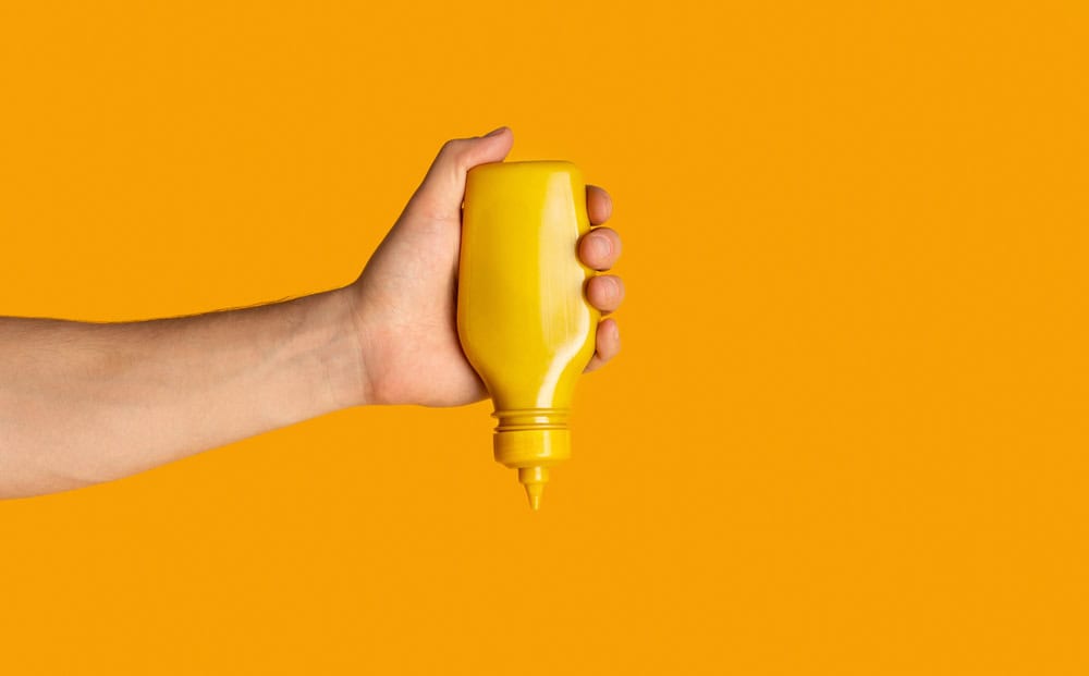 Male hand squeezing bottle with mustard on orange background, close up. Space for text