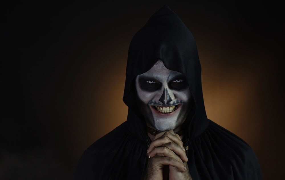 Creepy, crazy man with skeleton makeup in hood. Guy executioner making angry faces, shaking head, trying to scare. Voodoo rituals. Baron Saturday. Halloween party. Black background