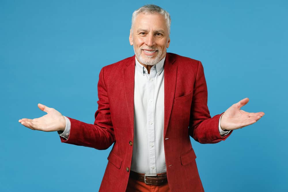 Smirked smiling confused elderly gray-haired mustache bearded business man wearing red jacket suit standing spreading hands looking camera isolated on blue color wall background studio portrait