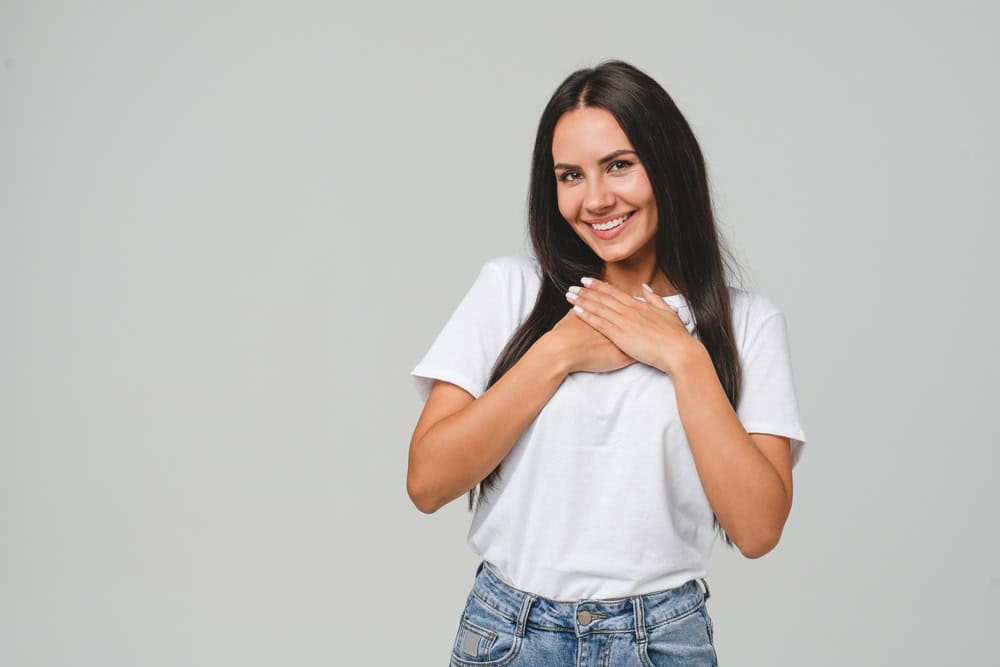 Touched pleased with a compliment cheerful happy caucasian young woman in white T-shirt isolated in grey background. Praise recommendation, sweet heart