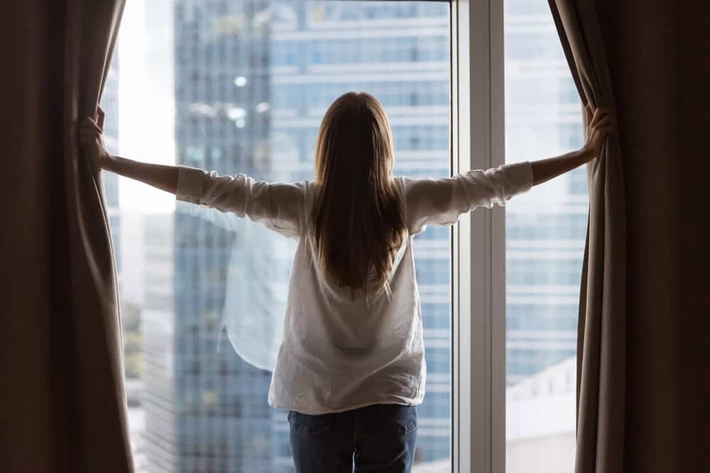 Rear view of young homeowner woman opening drapes at big window glass in new rented apartment, modern real estate property, enjoying city urban view, parting curtains