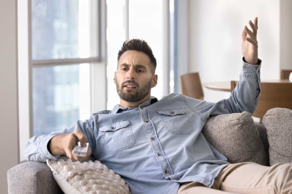 Nervous young Latin man using TV remote control on home couch, feeling annoyed, angry, concerned, watching football match, show, getting problems with broadcasting