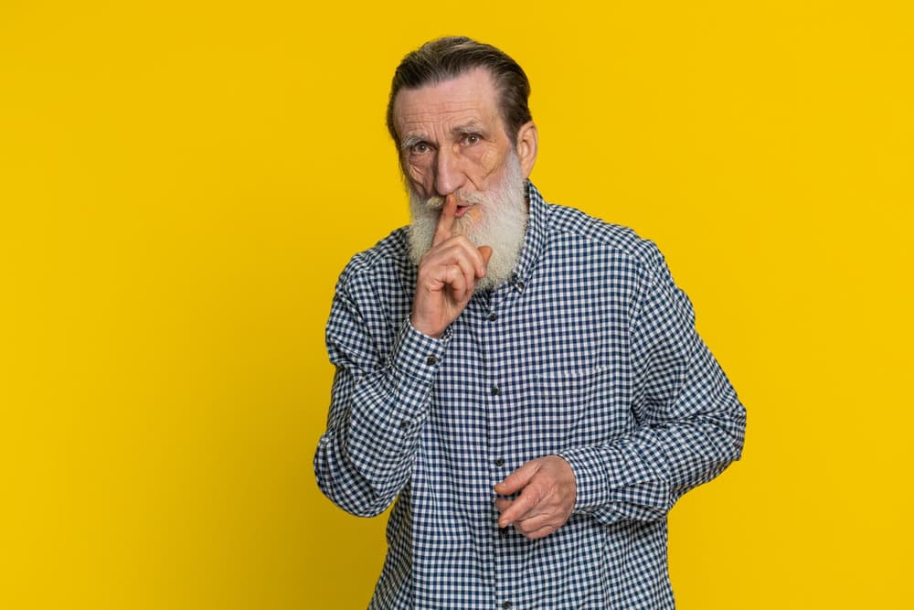 Shh be quiet please. Senior old man presses index finger to lips makes silence gesture sign do not tells secret stop talk gossip confidential privacy. Elderly bearded grandfather on yellow background
