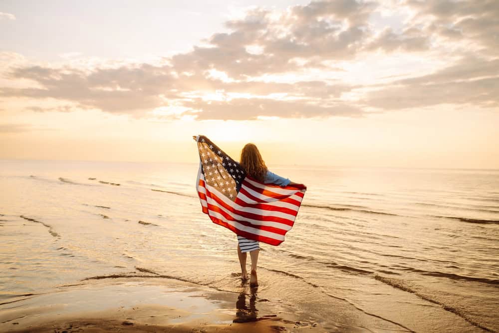 Happy woman with an American flag having fun on the seashore. Young tourist feeling free and celebrating American Independence Day. Concept of 4th of July, relaxation, enjoyment.
