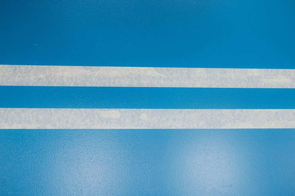 Two parallel strips of masking tape are applied to a blue epoxy floor coating, creating clean lines for a precise finish
