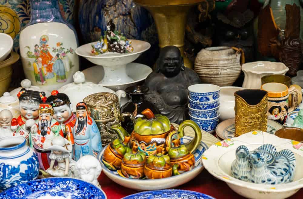 A diverse collection of antique and vintage ceramics, porcelain, and pottery on display at a flea market or antique shop in Bandung, Indonesia. A treasure trove of old figurines, teapots, and plates.