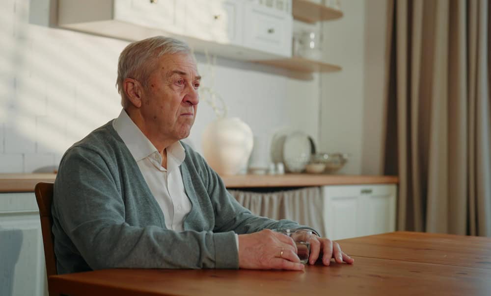 Widower sitting alone in home kitchen, missing and remembering, portrait. Sad senior man drinking water or calming tea, health care in old age, psychological problems and emotions of grey-haired man