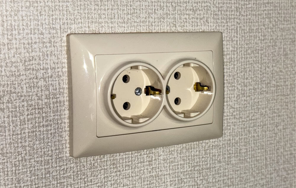 A wall shows an electrical outlet with two socket openings. The sockets are set into a plain surface, and there are two metal prongs visible in each opening.