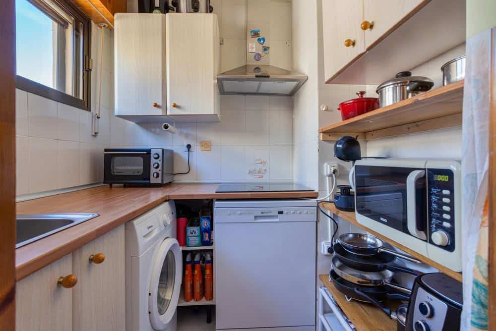 Alicante Spain 12 February 2026: Small apartment kitchen with dishwasher, washing machine, cooktop and countertop appliances arranged in compact Mediterranean residential interior.