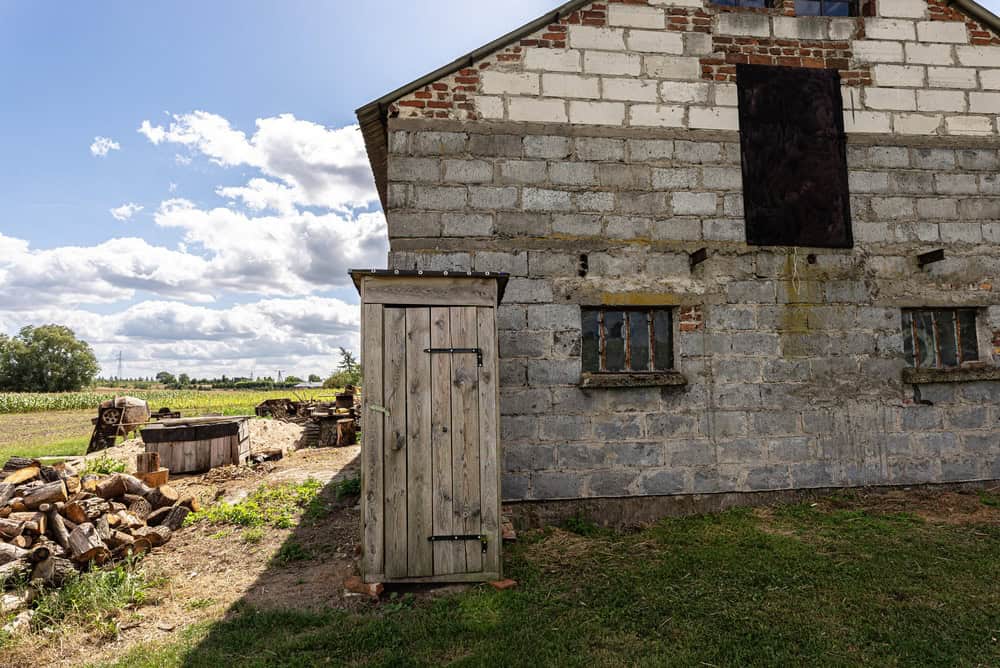 Wooden outdoor latrine standing by a farm building with a field in the background under a blue sky